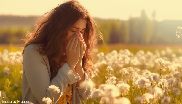 A woman blows her nose amidst a field of dandelions, illustrating the photic sneeze reflex triggered by bright visuals.